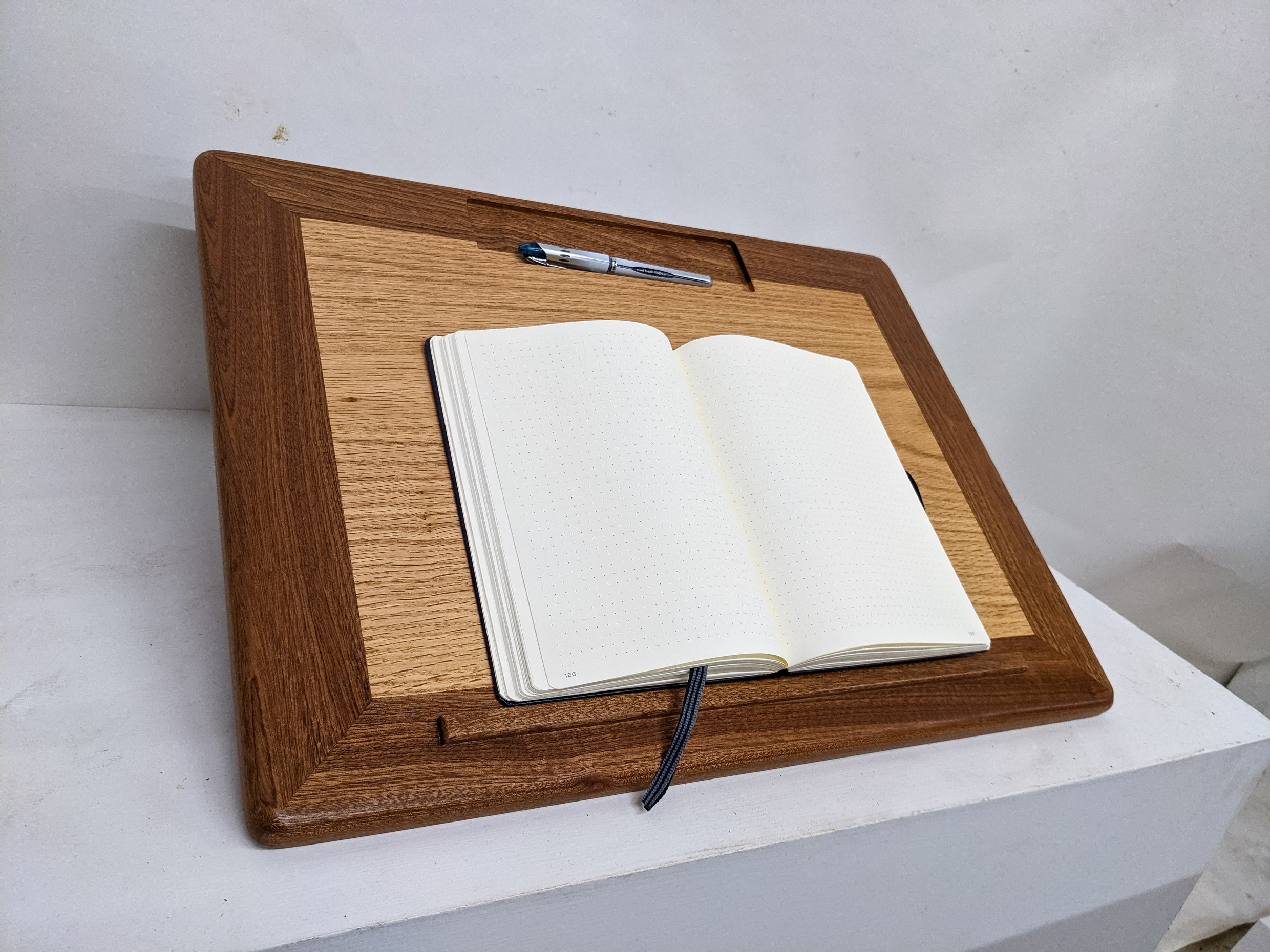 The white pages of a journal is exposed and open as the journal sits on the protruding mahogany lip of our Leather Cushion Lap Desk. The desk is framed in Mahogany and maintains an oak interior. At the top, a rectangular divet holds a pen.